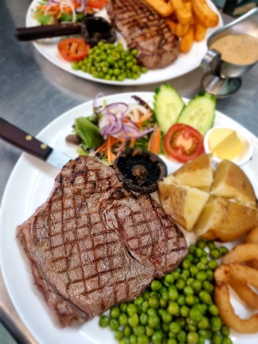 A grilled steak with a flat mushroom, tomato, peas and a jacket potato.